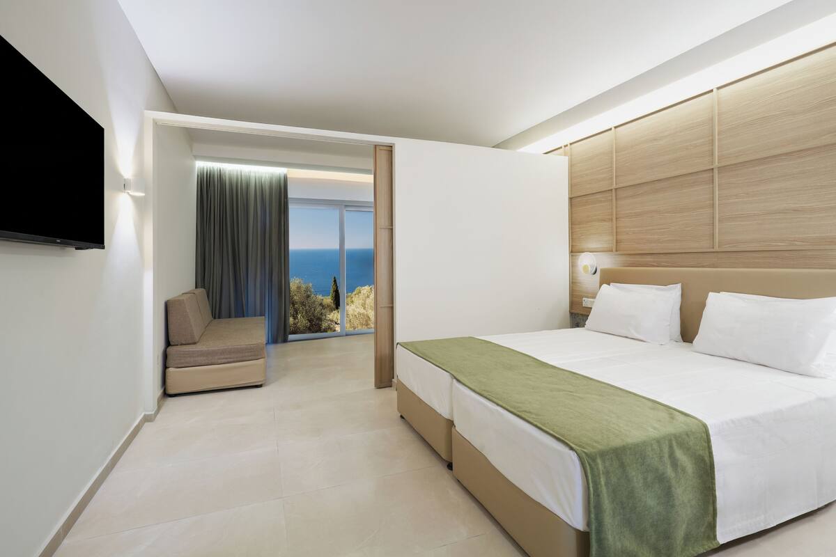 A modern hotel room with a large bed, a sofa, and a TV. There is a balcony with a view of the sea.