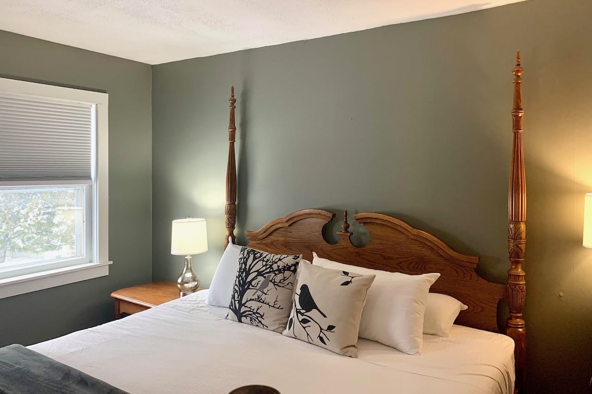 A bedroom with a large bed, a wooden headboard, two wall-mounted lamps, and a window with blinds.