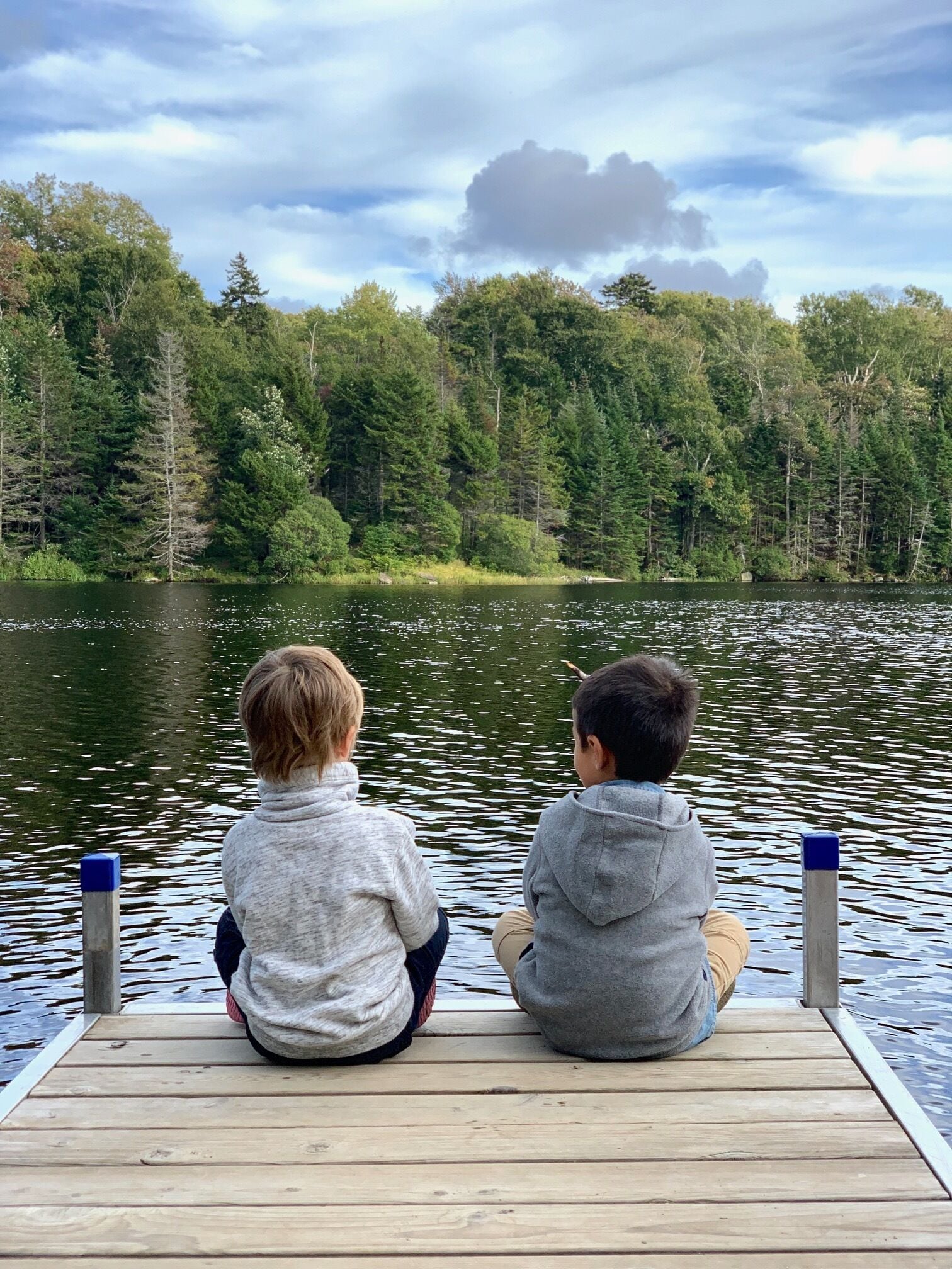 Two children sitting on a wooden dock overlooking a lake surrounded by dense forest.