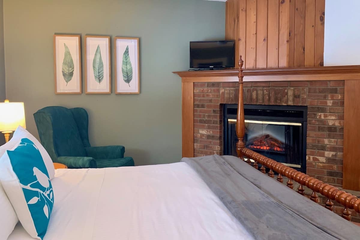 A bedroom with a bed, a green armchair, a fireplace, and three framed leaf artworks on the wall.