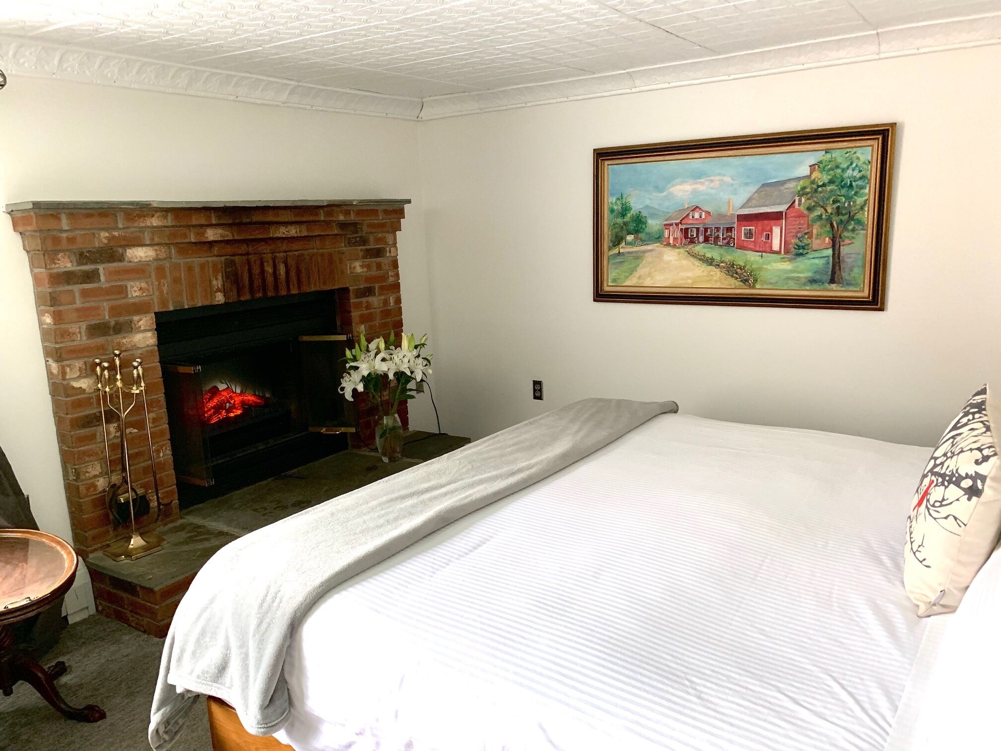 Un dormitorio con chimenea de ladrillo, un cuadro de paisaje rural y una cama con edredón blanco.