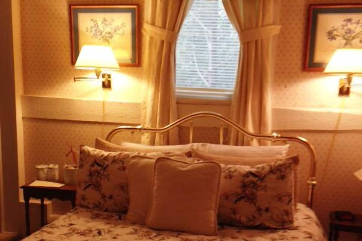 A neatly made bed with floral bedding, a window with curtains, and two wall-mounted lamps.