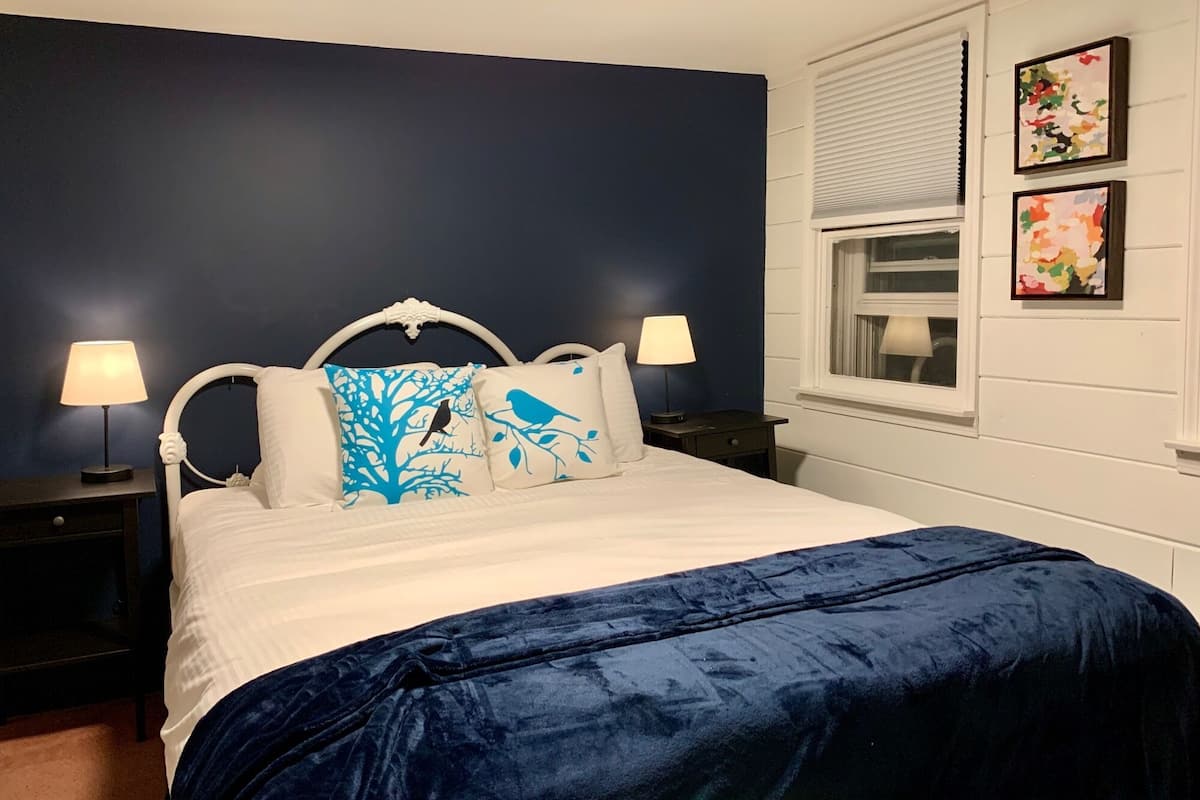 A bedroom with a dark blue wall, a white bed with a blue and white patterned headboard, two bedside tables with lamps, and a window with blinds.