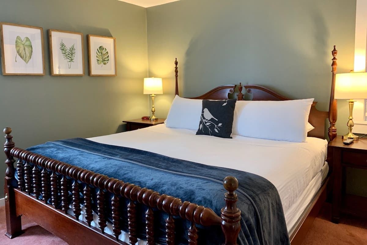 A neatly made bed with a wooden headboard, a blue throw blanket, and two bedside lamps. There are framed botanical prints on the wall.