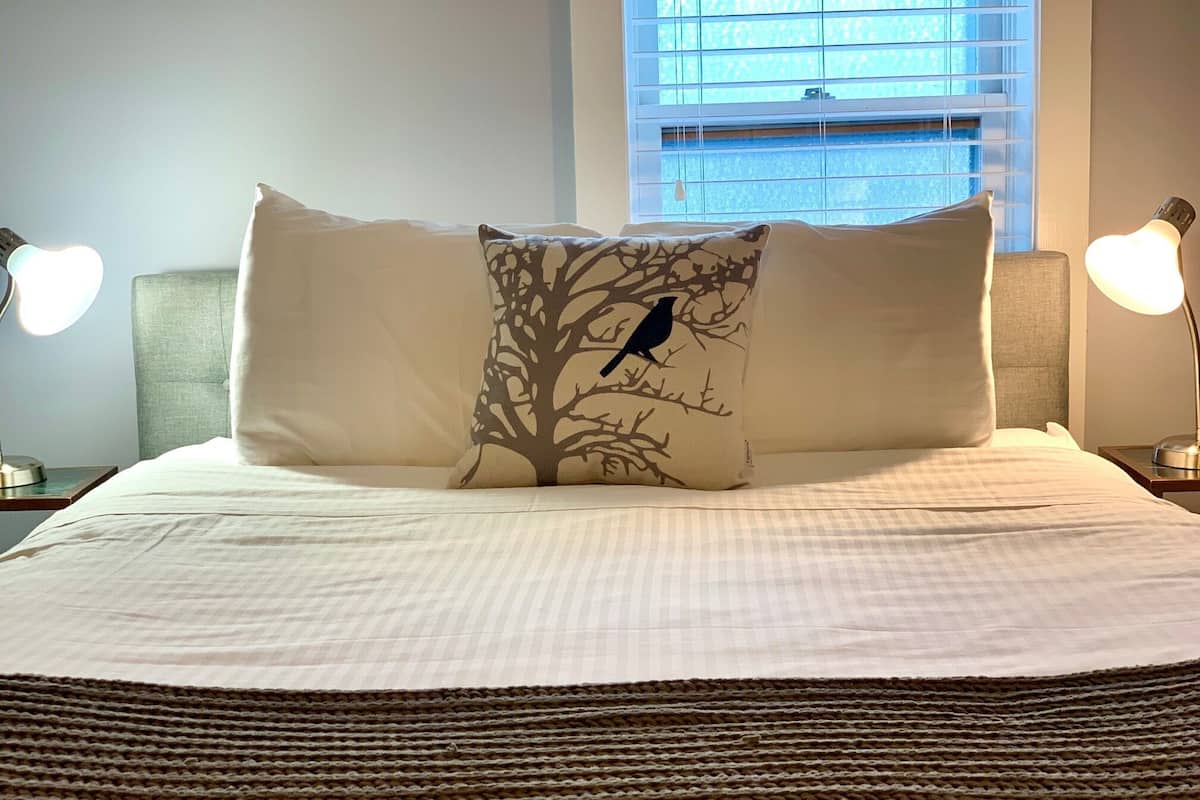 A neatly made bed with a tree branch pillow, two bedside lamps, and a window with blinds.