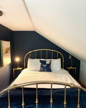 A bedroom with a sloped ceiling, a bed with a metal frame, white bedding, and a decorative pillow. There are two bedside tables with lamps. - The Nutmeg (Wilmington)