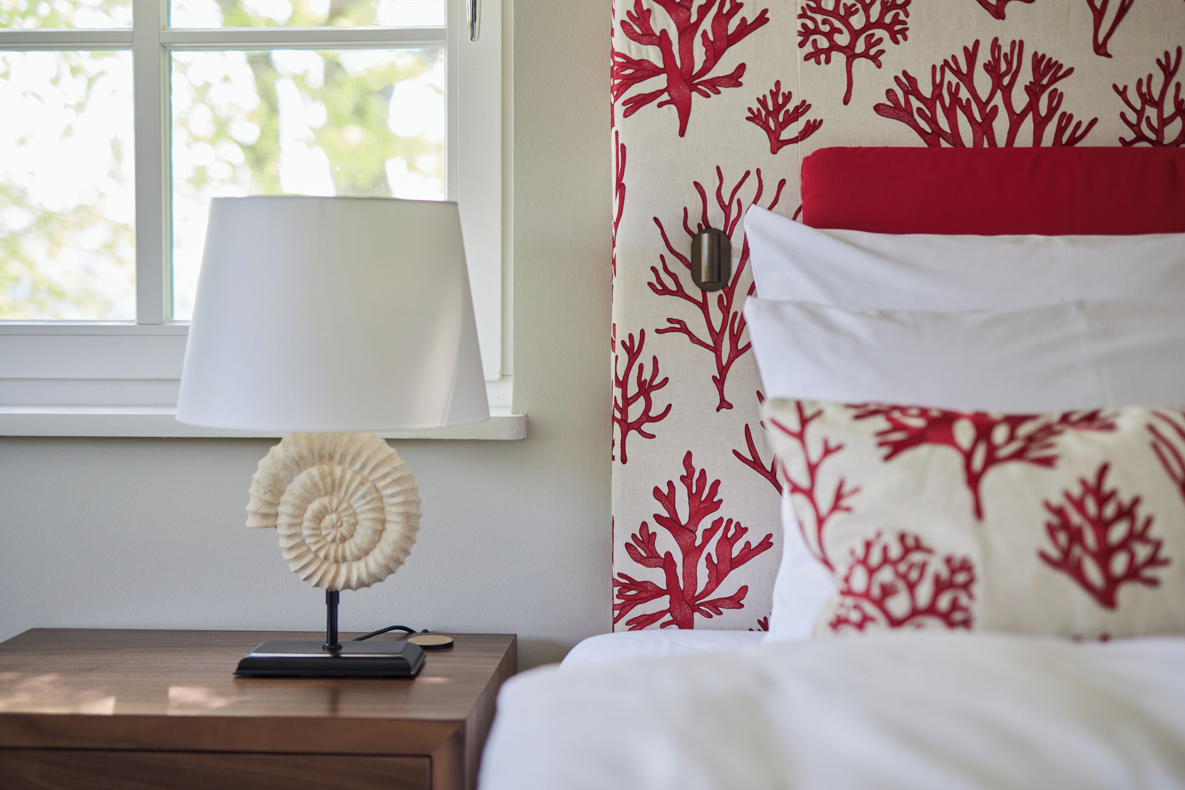 Een net opgemaakt bed met een rood-wit gebeddeken, een wit kussen en een witte lamp op een houten nachtkastje.