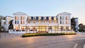 Un hotel grande y blanco de estilo arquitectónico clásico, con una entrada central y un vestíbulo cubierto, y un jardín bien cuidado.
