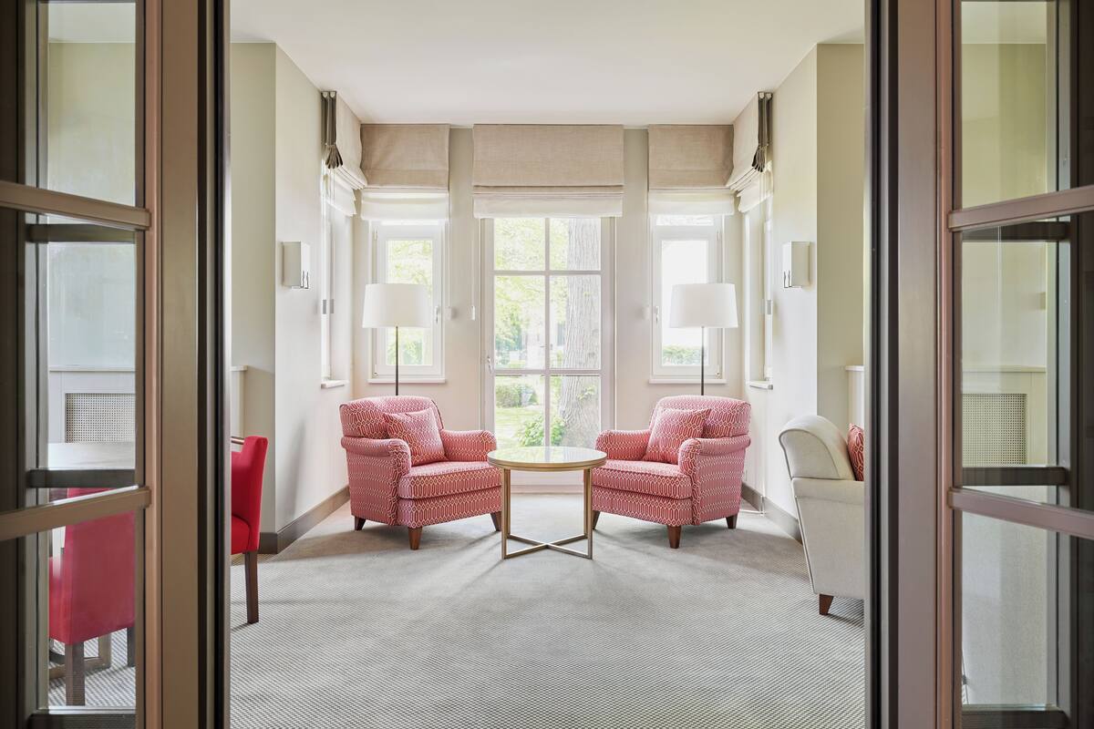 Ein Zimmer mit zwei rot gemusterten Sesseln, die einem runden Tisch gegenüberliegen, einem weißen Sessel und einem großen Fenster mit Jalousien.
