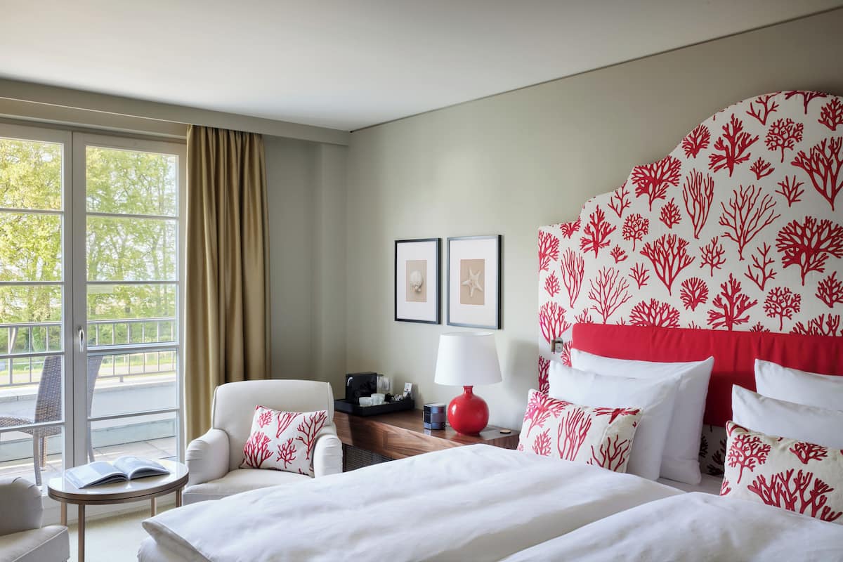 Ein Hotelzimmer mit einem großen Bett, einer Sitzecke mit einem Sessel und einem kleinen Tisch, einem Fenster mit Vorhängen und einer Wand mit korallenmusteriertem Wandpapier.