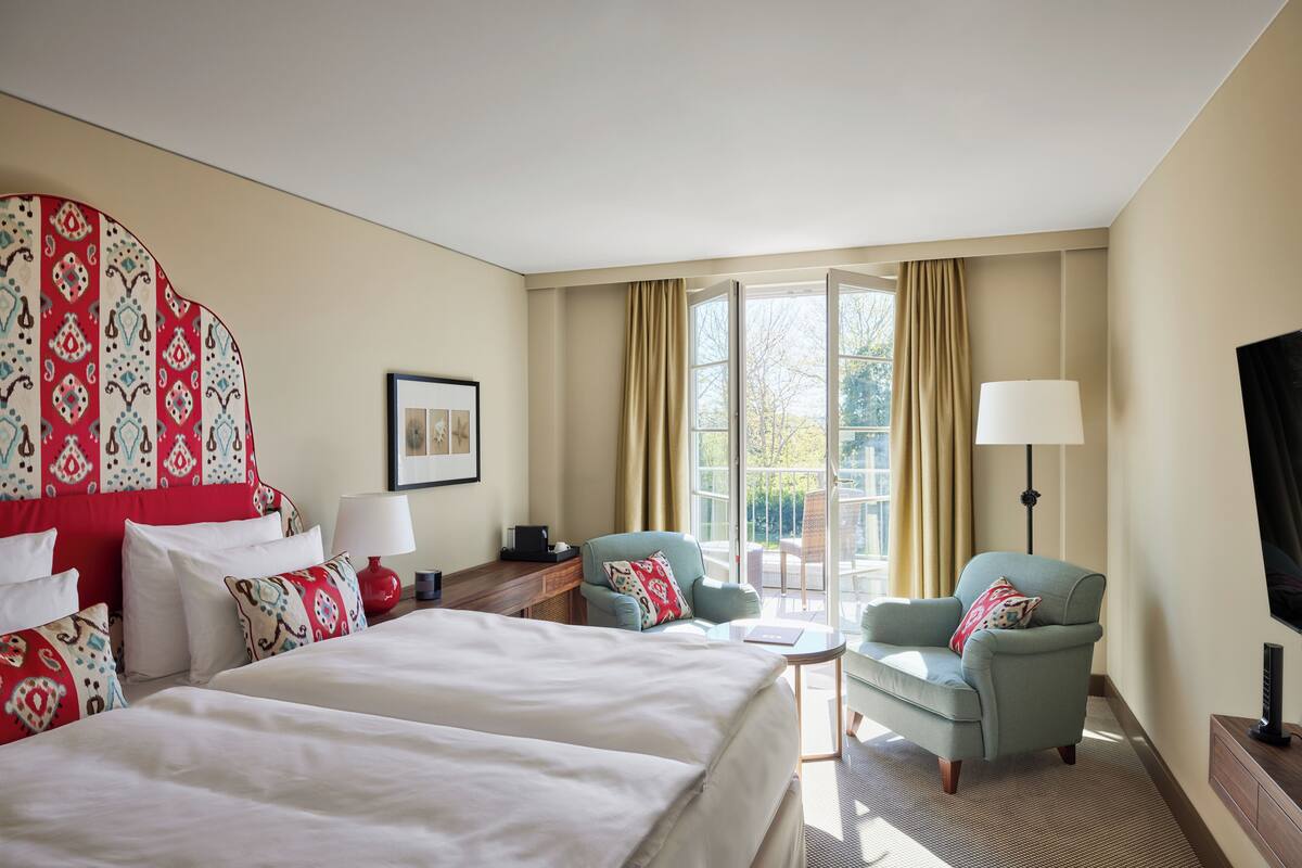 Ein Hotelzimmer mit zwei Betten, einer Sitzecke mit Sessel und Tisch, einem Fernseher und einem großen Fenster mit Vorhängen.