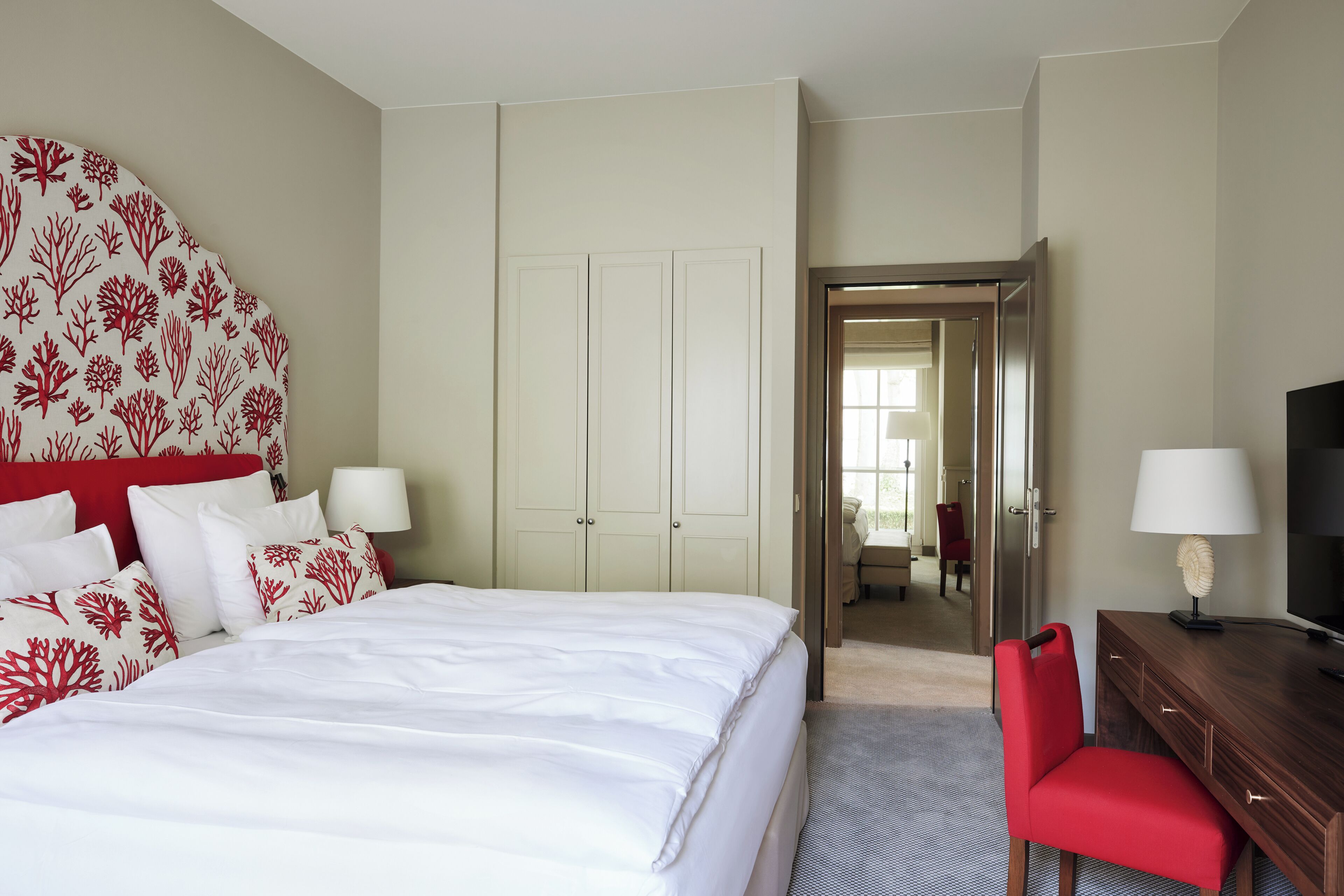 A hotel room with a bed, a red chair, a wooden dresser, a TV, and a door leading to another room.