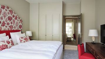 A hotel room with a bed, a red chair, a wooden dresser, a TV, and a door leading to another room.