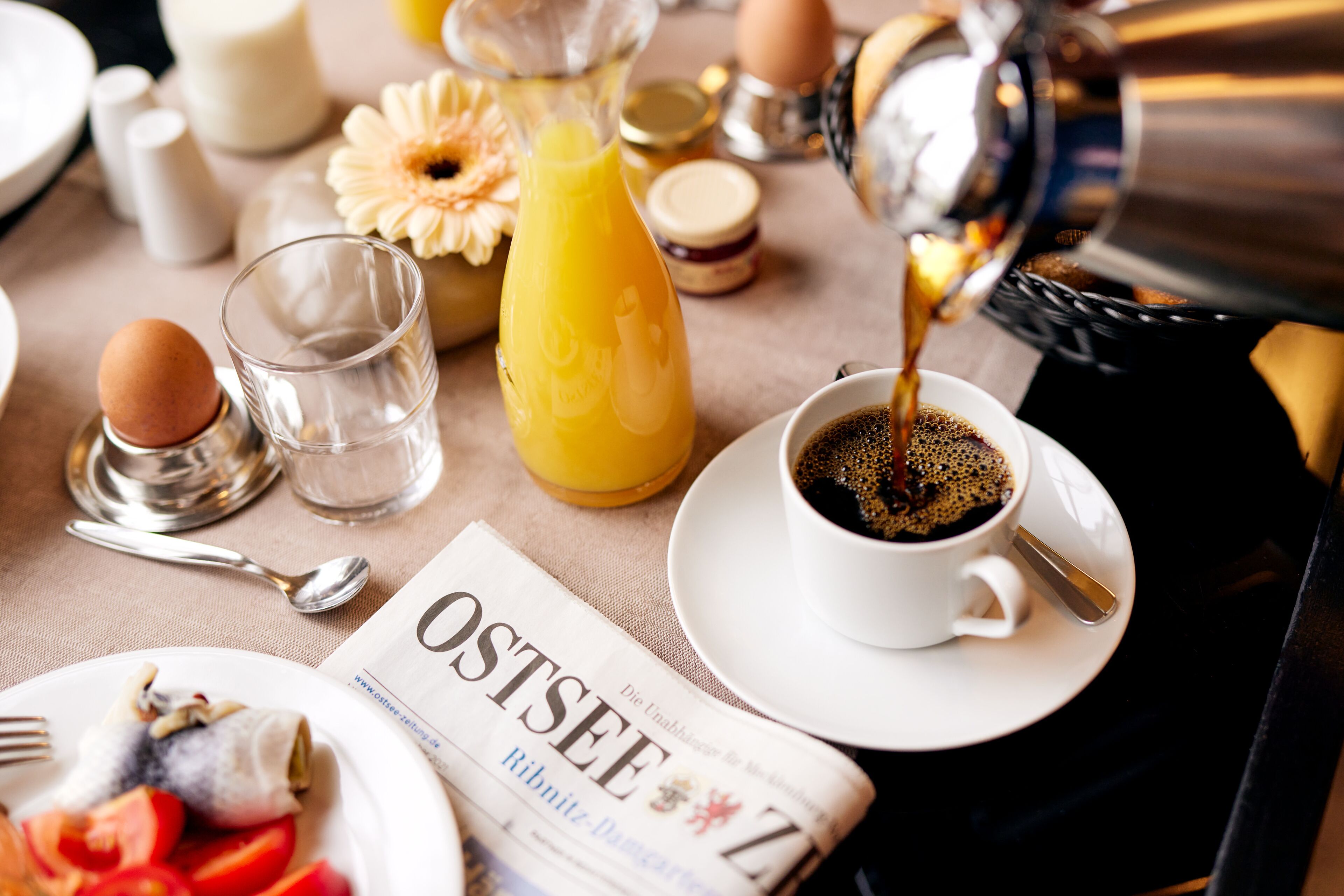 Une table de petit-déjeuner avec un journal, une tasse de café, un verre de jus d’orange et une assiette contenant des œufs et des tomates.