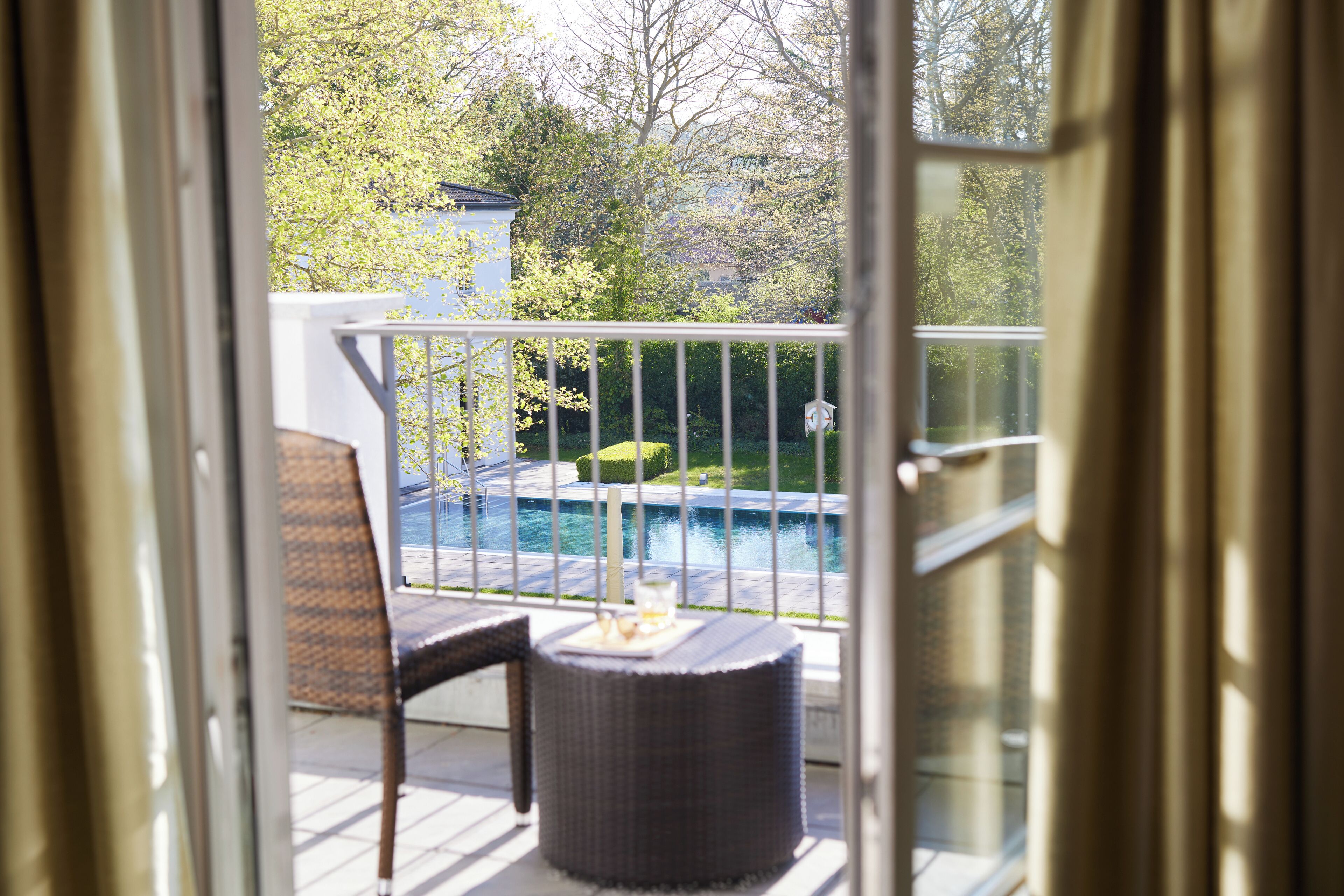 Ein Balkon mit Korbmöbeln und einem kleinen Tisch, mit Blick auf einen Swimmingpool und einen Garten.