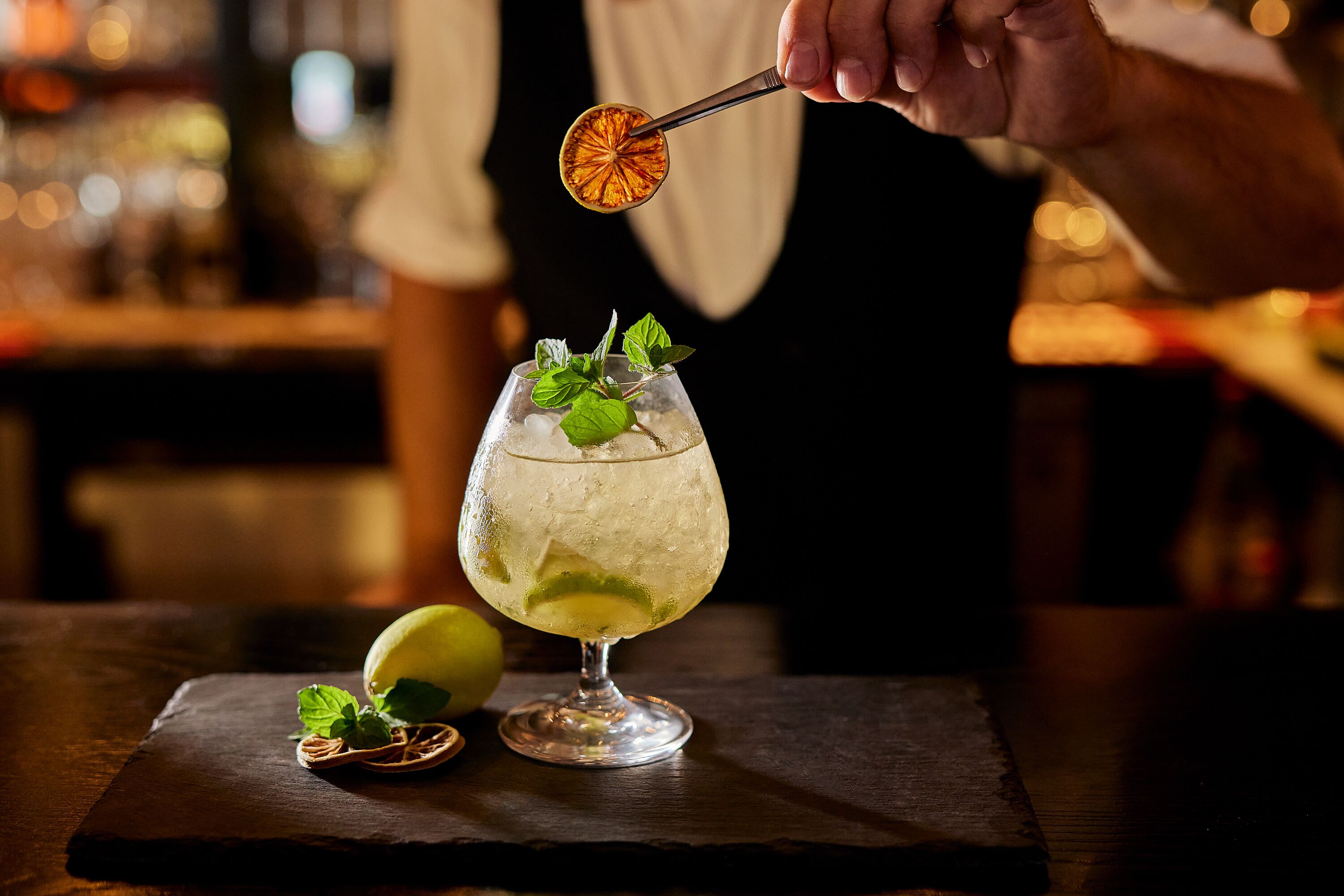 A cocktail glass with ice, lime, and mint leaves on a dark slate coaster.