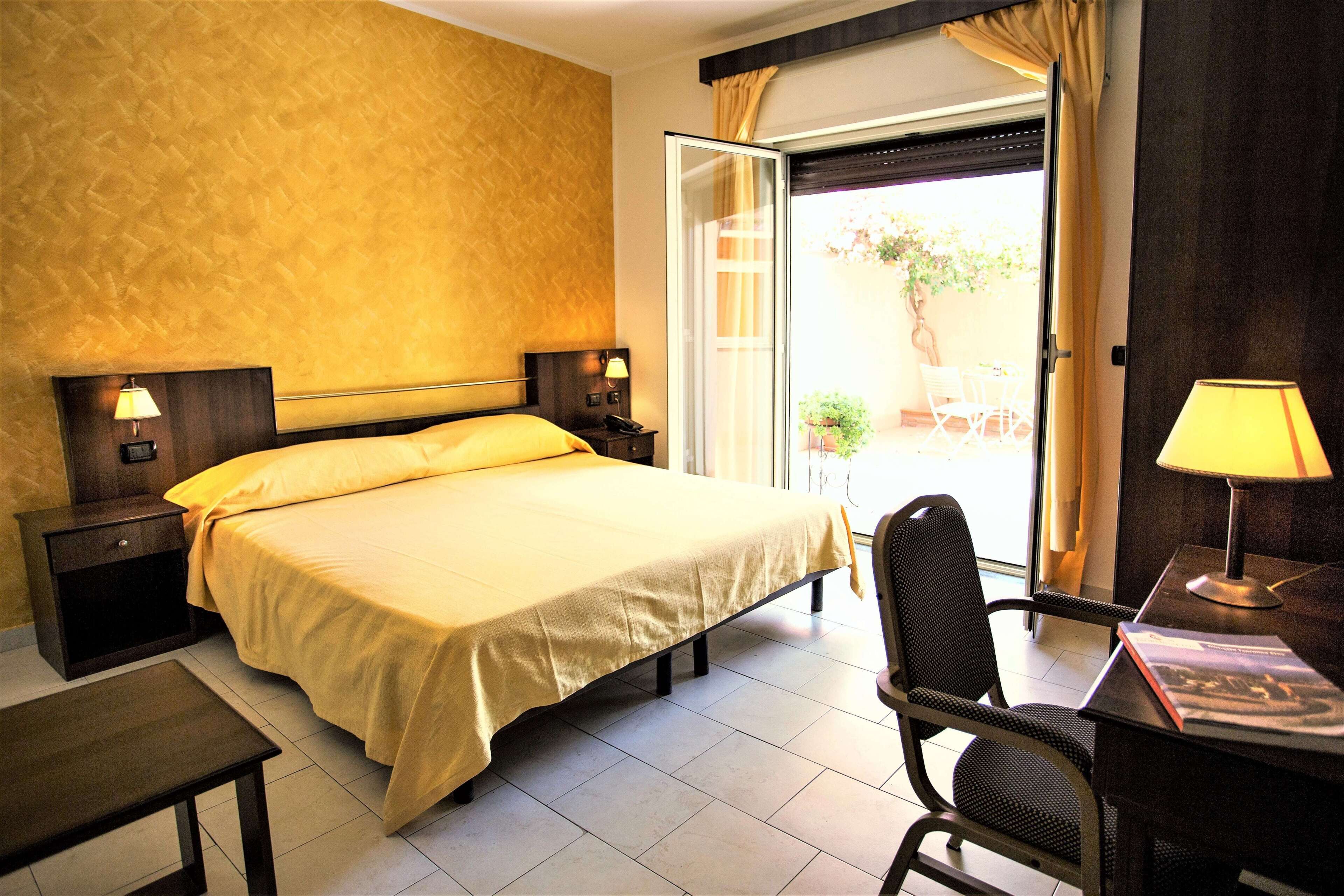 A hotel room with a bed, bedside tables, a desk with a chair, and a window with curtains.