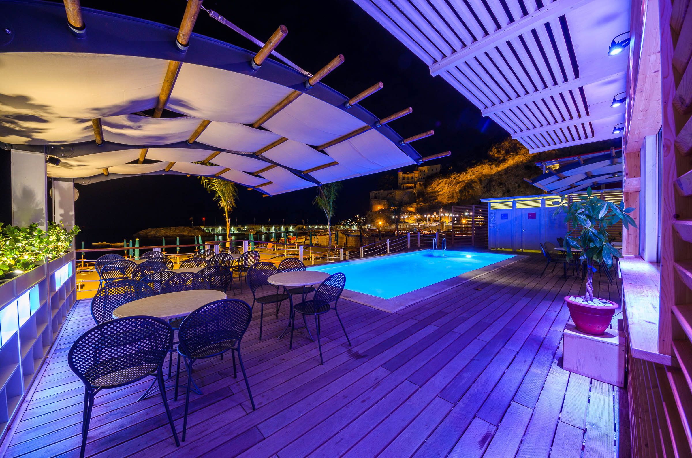 An outdoor pool area with wooden decking, metal chairs, and a covered seating area.