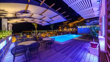 An outdoor pool area with wooden decking, metal chairs, and a covered seating area.