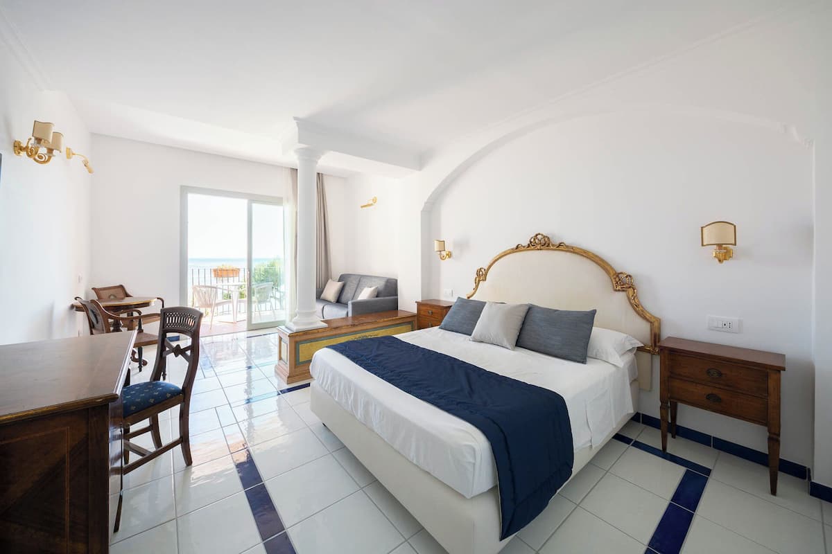 A bedroom with a large bed, a desk, and a chair. There is a balcony with a view.