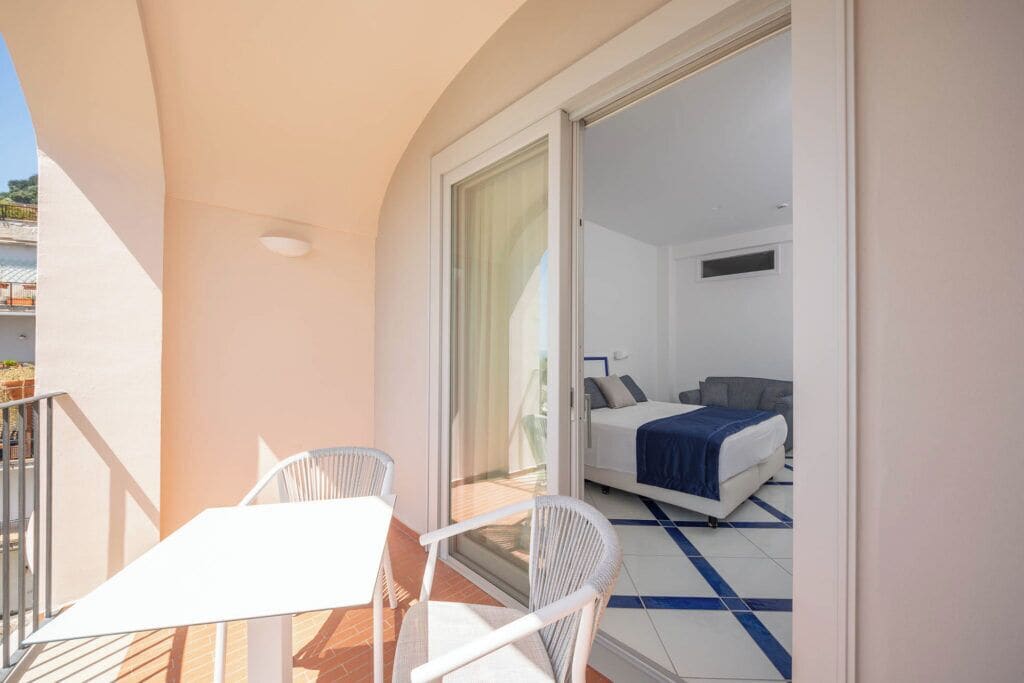 A balcony with a white table and chairs, a bed with a blue blanket, and a sliding glass door leading to a bedroom.