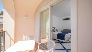 A balcony with a white table and chairs, a bed with a blue blanket, and a sliding glass door leading to a bedroom.