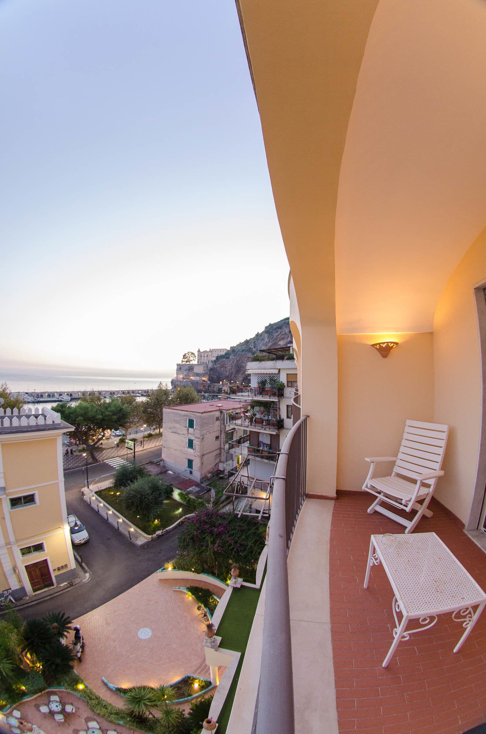 Ein Balkon mit weißem Stuhl, einem kleinen Tisch und Blick auf das Meer und die Gebäude.