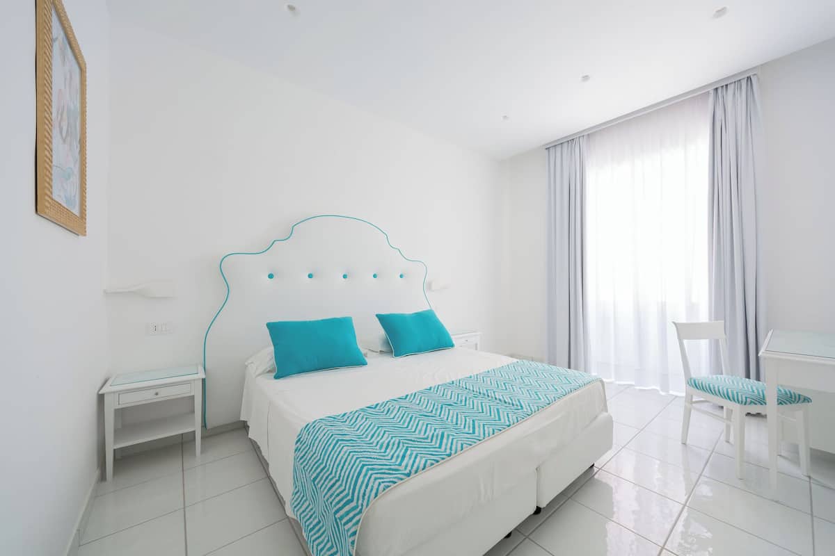 A modern bedroom with a white bed, two turquoise pillows, a white nightstand, a white chair, and a large window with curtains.