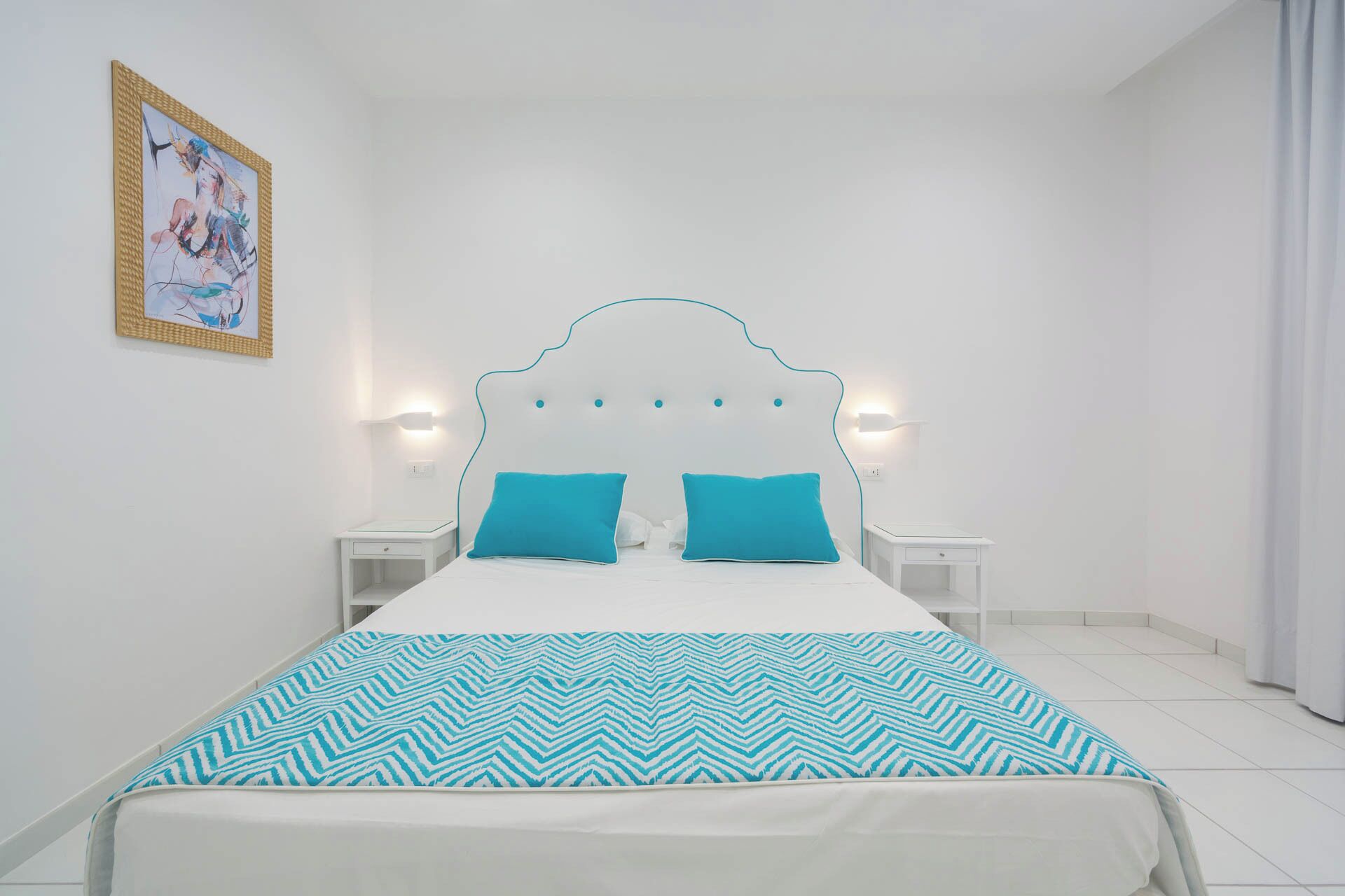 Un dormitorio con una cama blanca, dos almohadas turquesas, un cabecero blanco y un cuadro colgado en la pared.