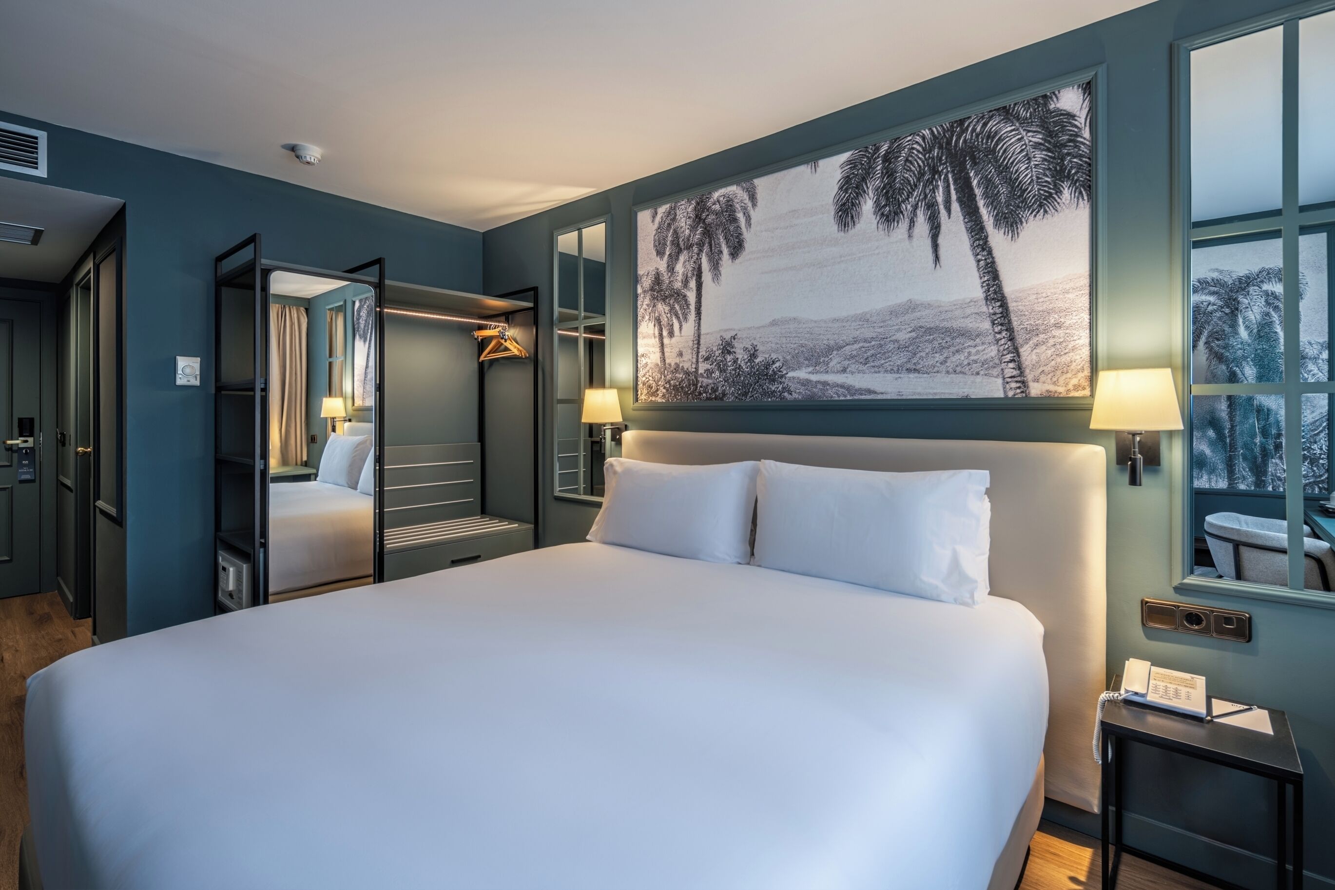 Quarto de hotel com uma cama grande, uma pintura de palmeiras, um espelho e uma mesa de cabeceira com abajur.