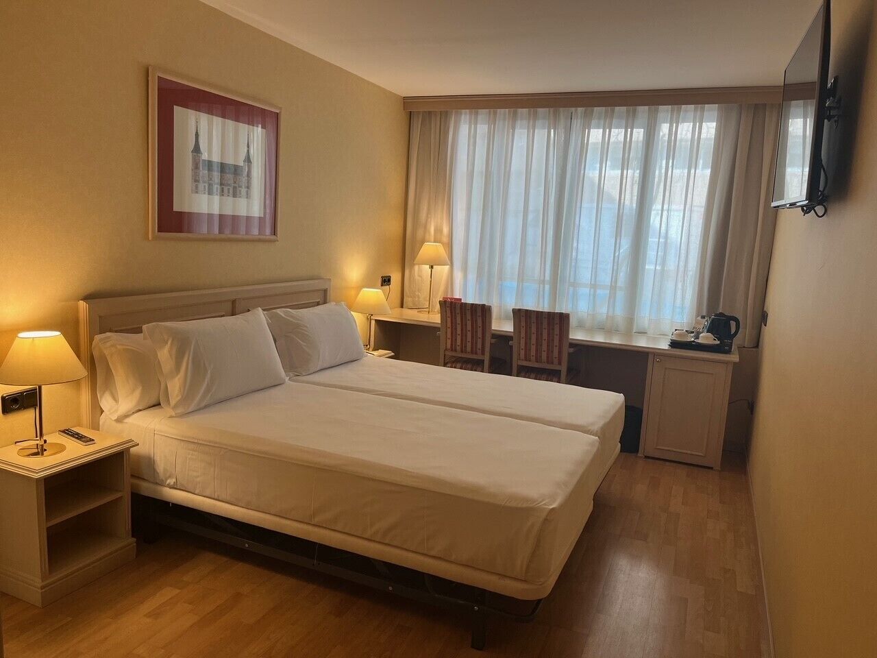 A hotel room with a double bed, bedside tables with lamps, a small desk with a chair, a window with sheer curtains, and a framed picture on the wall.