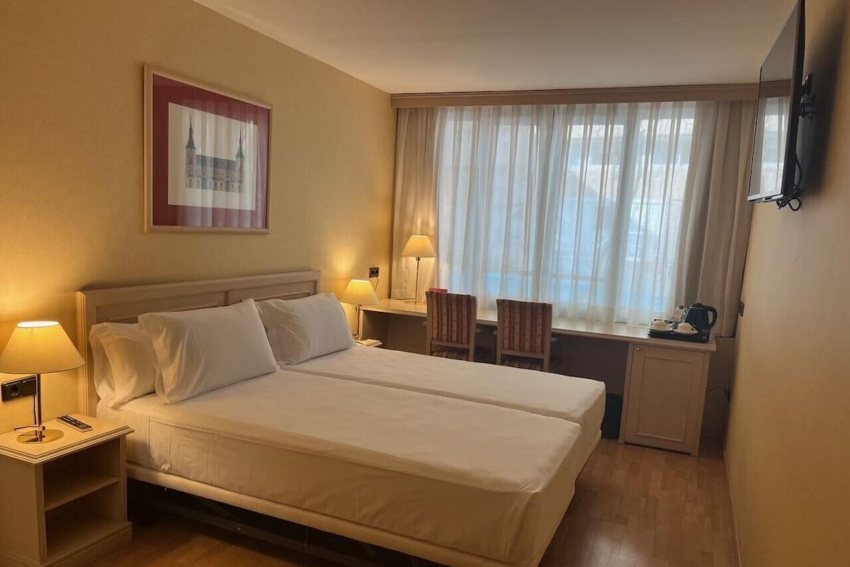 A hotel room with a double bed, bedside tables with lamps, a small desk with a chair, a window with sheer curtains, and a framed picture on the wall.