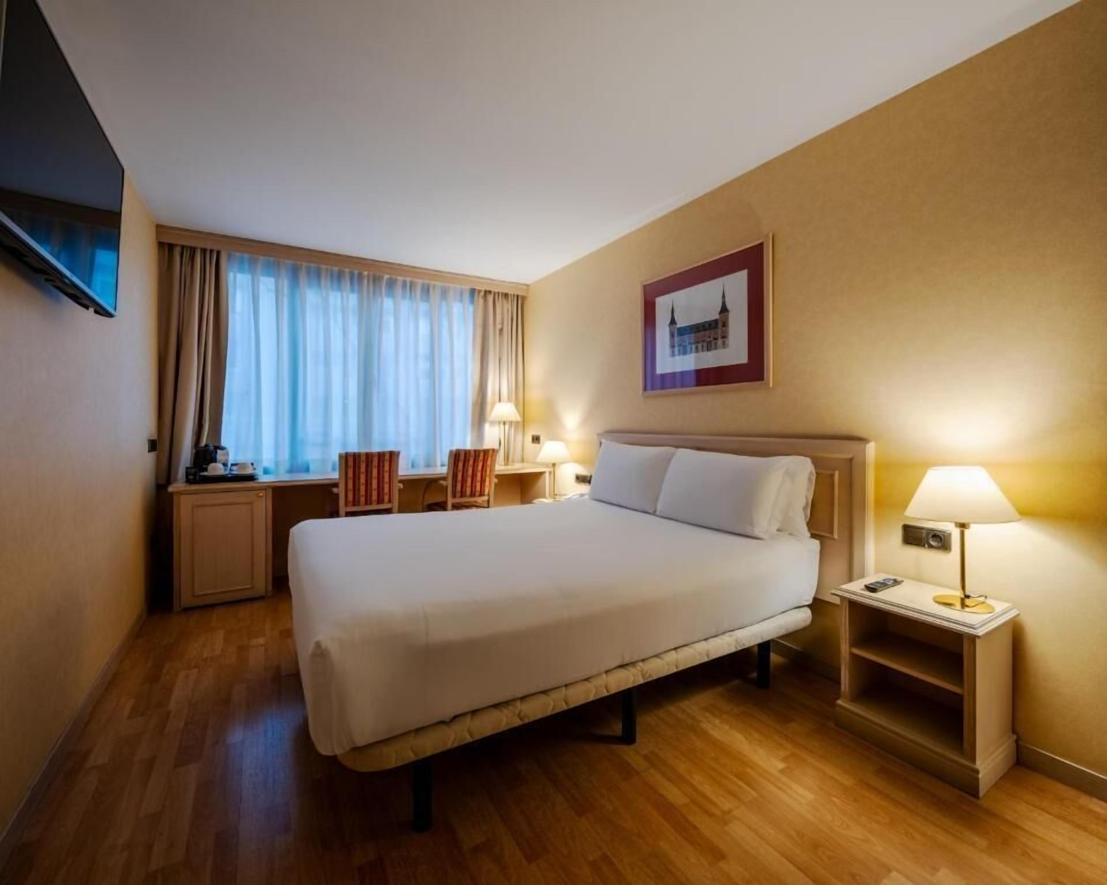 Quarto de hotel com cama, mesas de cabeceira, escrivaninha, cadeira, televisão e janela com cortinas.