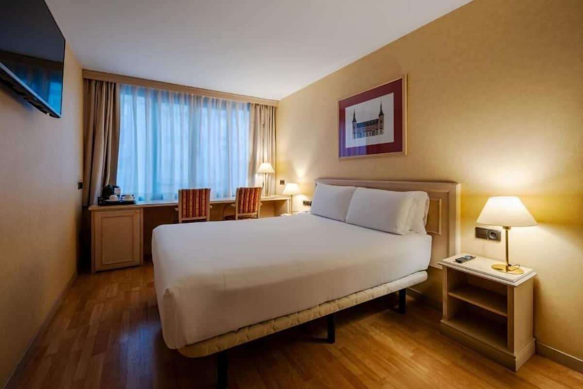 Quarto de hotel com cama, mesas de cabeceira, escrivaninha, cadeira, televisão e janela com cortinas.