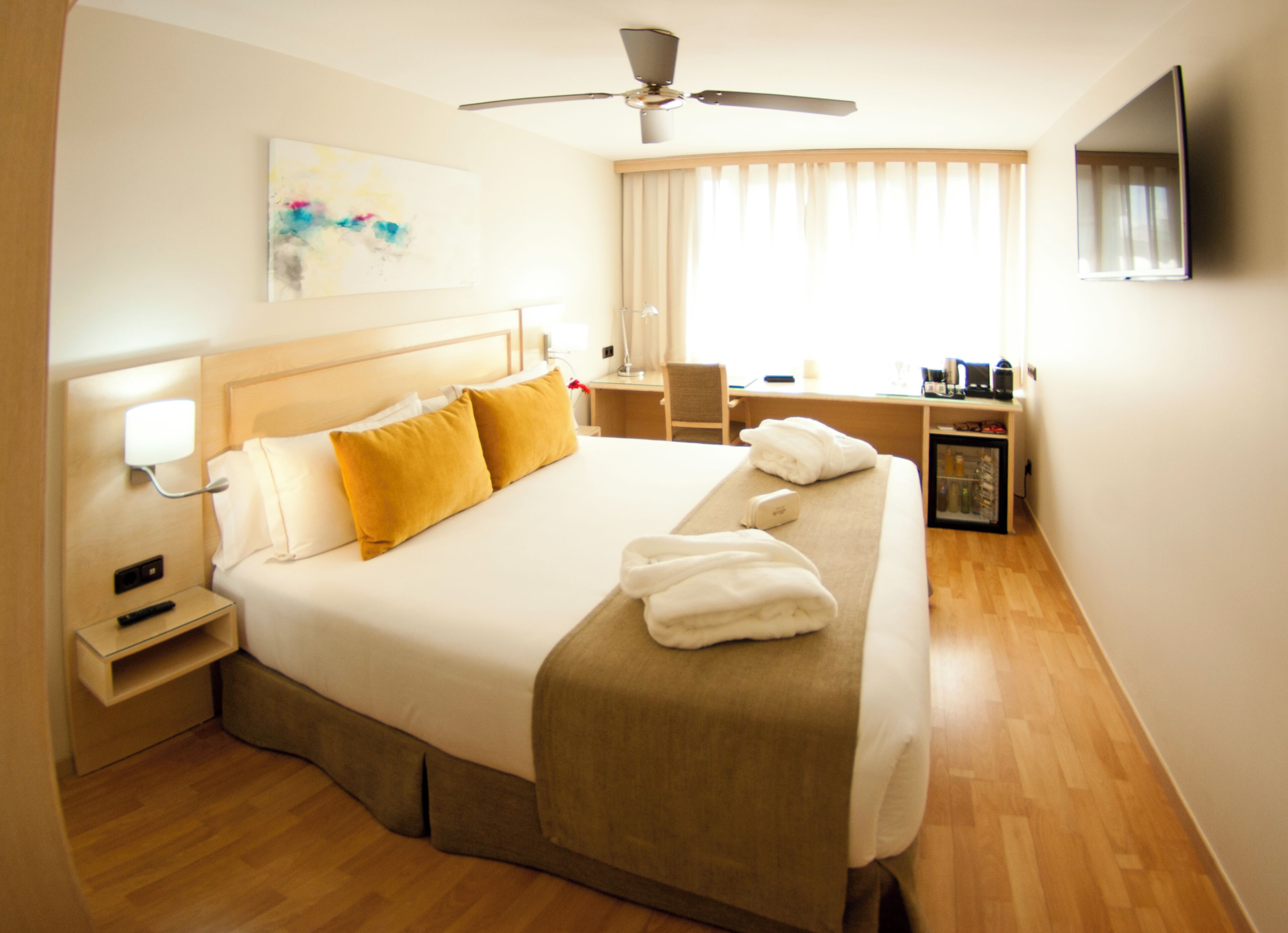 Quarto de hotel com uma cama grande, uma escrivaninha, uma cadeira, uma televisão e um ventilador de teto.