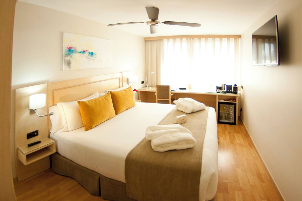 Quarto de hotel com uma cama grande, uma escrivaninha, uma cadeira, uma televisão e um ventilador de teto.