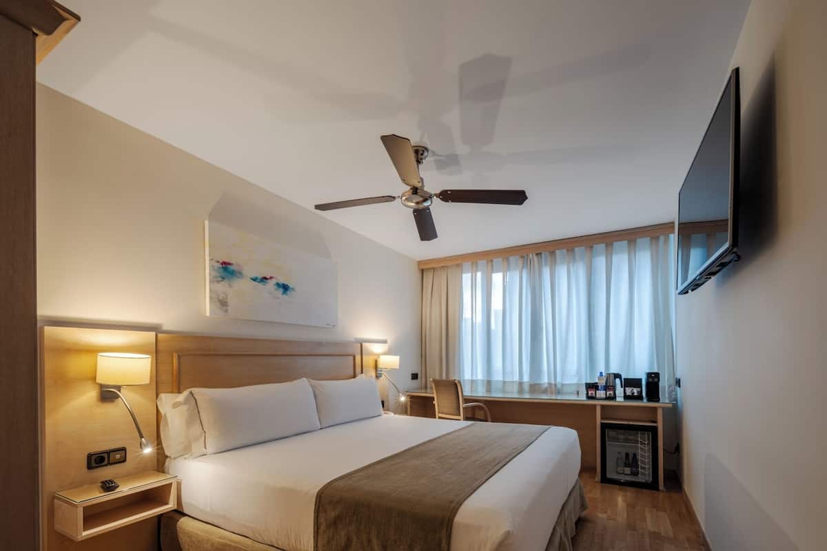 A hotel room with a bed, bedside tables, a desk, a chair, a television, and a ceiling fan.