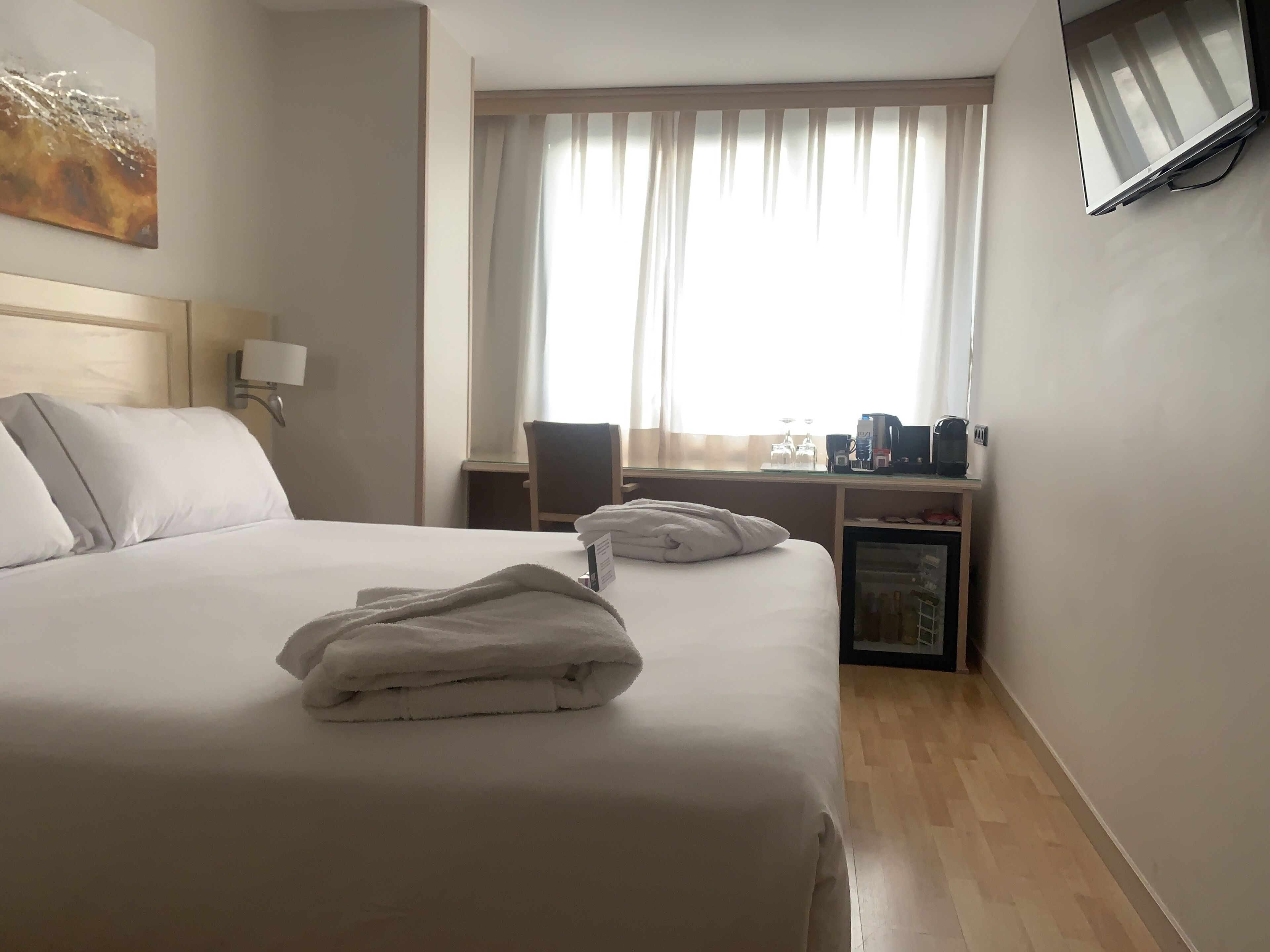 A hotel room with a bed, a desk, a chair, a TV mounted on the wall, and a window with curtains.