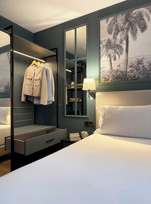 A hotel room with a bed, a nightstand, a bedside table, a wardrobe, a mirror, and a wall with a palm tree painting. - BYPILLOW Castellana (Madrid)