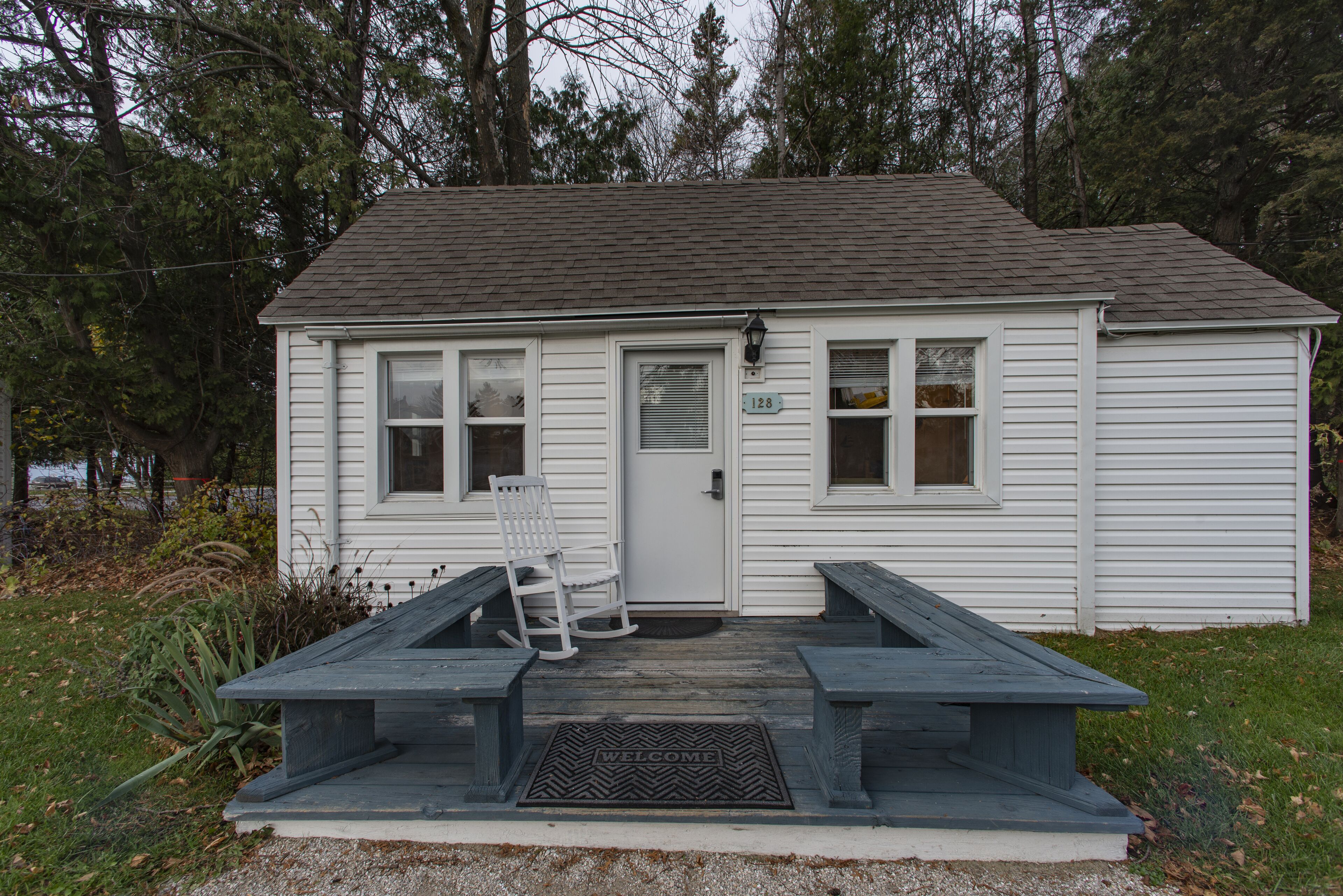 A small white building with a porch, a rocking chair, and a welcome mat.