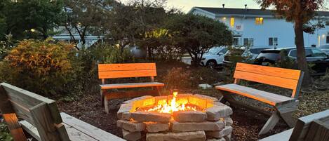 A fire pit with benches around it, surrounded by trees and a building in the background.