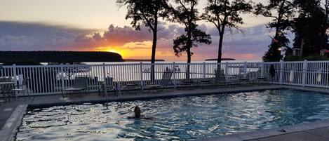 Una piscina con vista al atardecer, rodeada de sillas de descanso y una valla blanca.