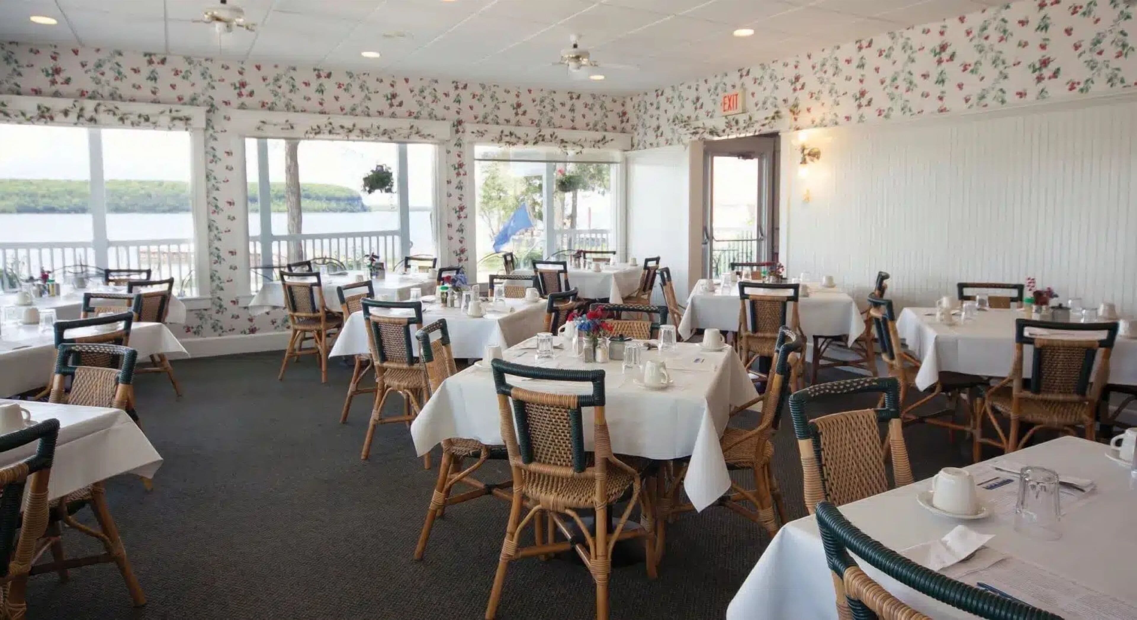A dining area with tables set for service, wicker chairs, and large windows offering a view of the outdoors.