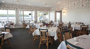 A dining area with tables set for service, wicker chairs, and large windows offering a view of the outdoors.