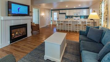 Una sala de estar con chimenea, televisor, sofá y una cocina al fondo.