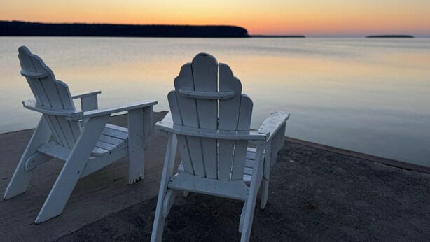 Dos sillas Adirondack blancas mirando un cuerpo de agua tranquilo con un atardecer de fondo.
