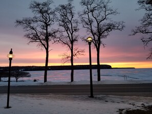 A winter scene with a sunset over a frozen lake, three street lamps, and bare trees.