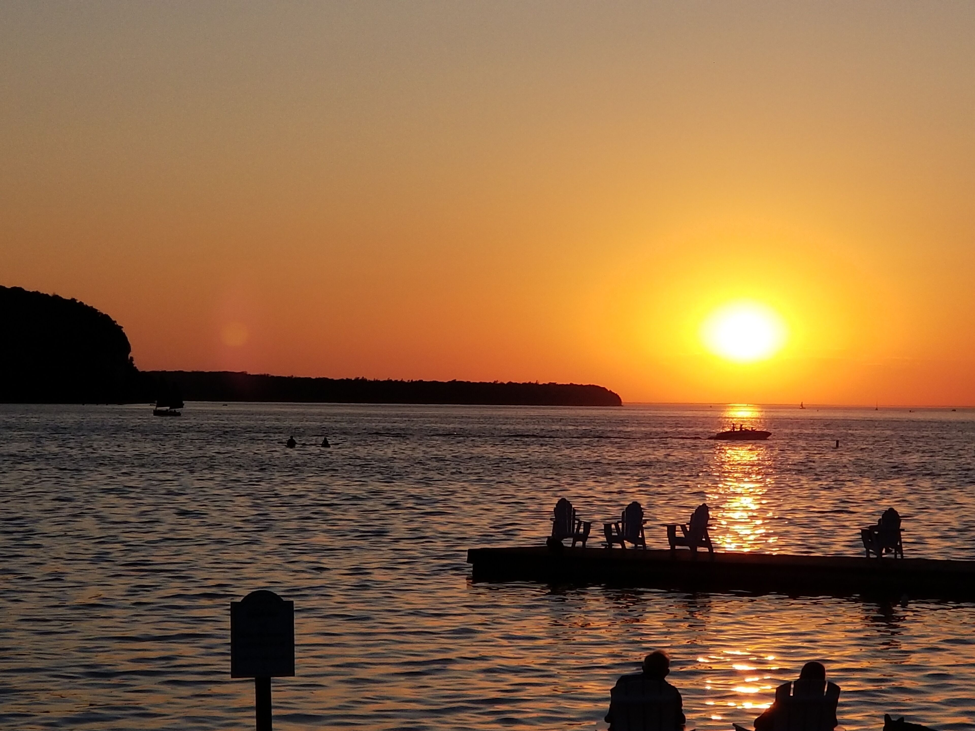 A sunset over a calm body of water with a pier and people sitting on chairs.