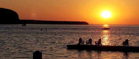 A sunset over a calm body of water with a pier and people sitting on chairs.