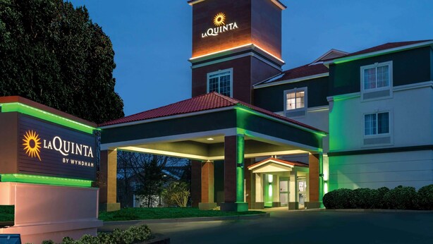 Un hotel con un letrero destacado que dice "LA QUINTA BY WYNDHAM", una entrada bien iluminada y una gran torre con el nombre del hotel.